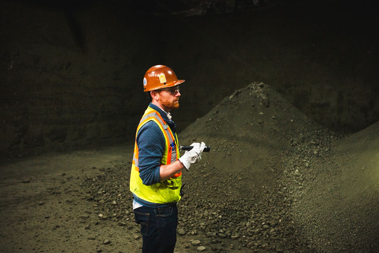 Hugo Staton, wearing an green vest and an orange hard hat, shines a flashlight on a pile of metal concentrate