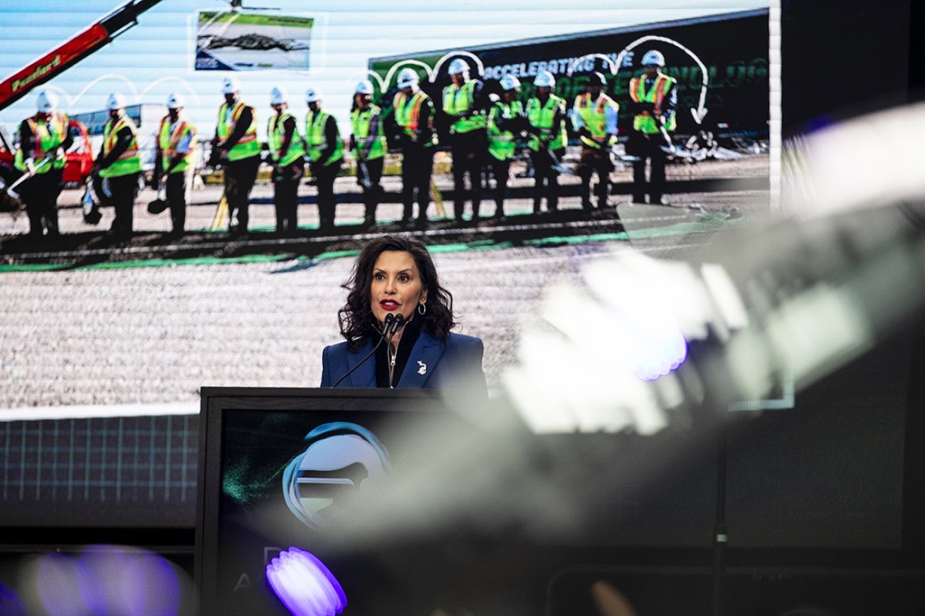 Gov. Gretchen Whitmer speaking on stage. Behind her is a photo of a groundbreaking.