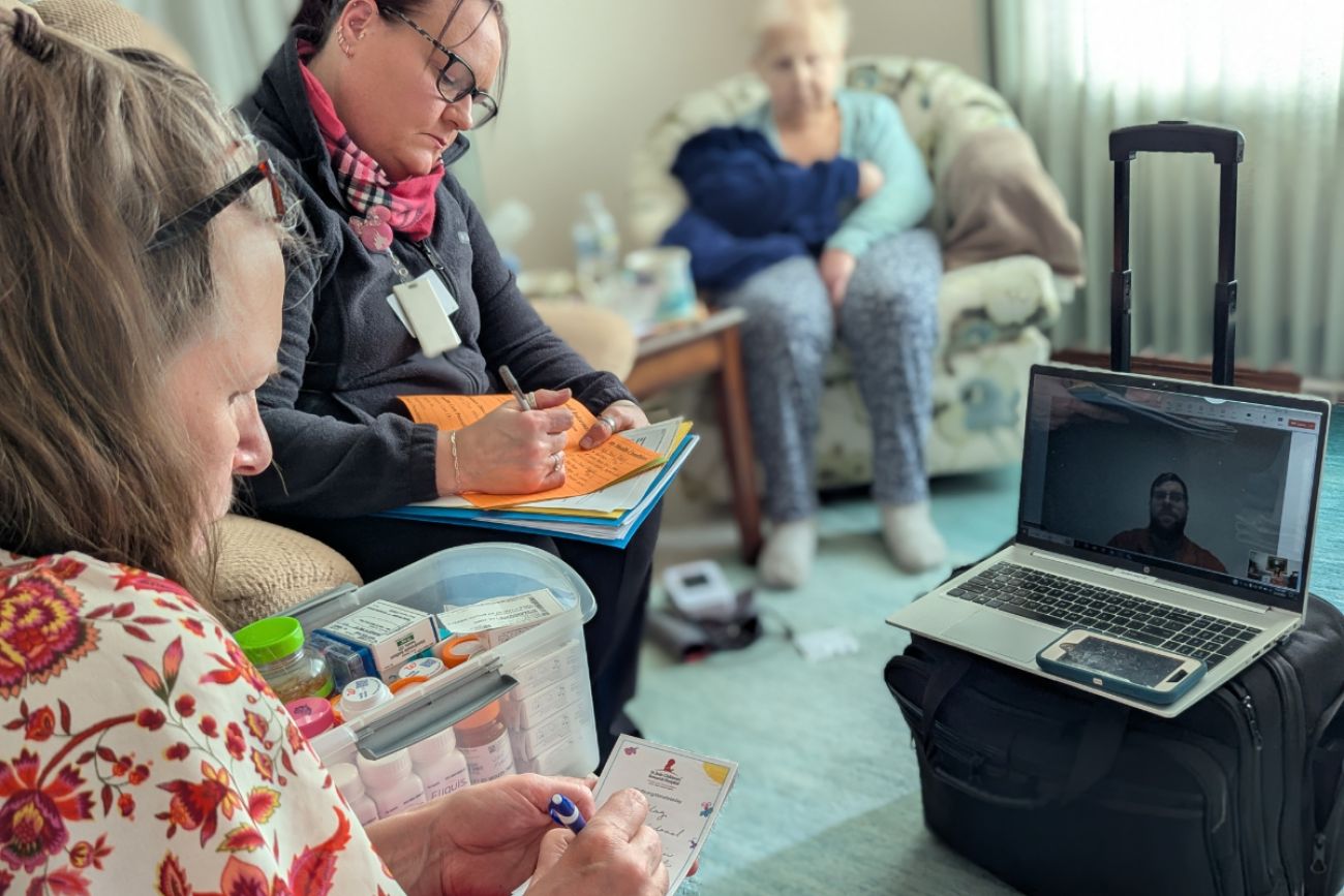 Two women take notes while taking to a pharmacist on a laptop.