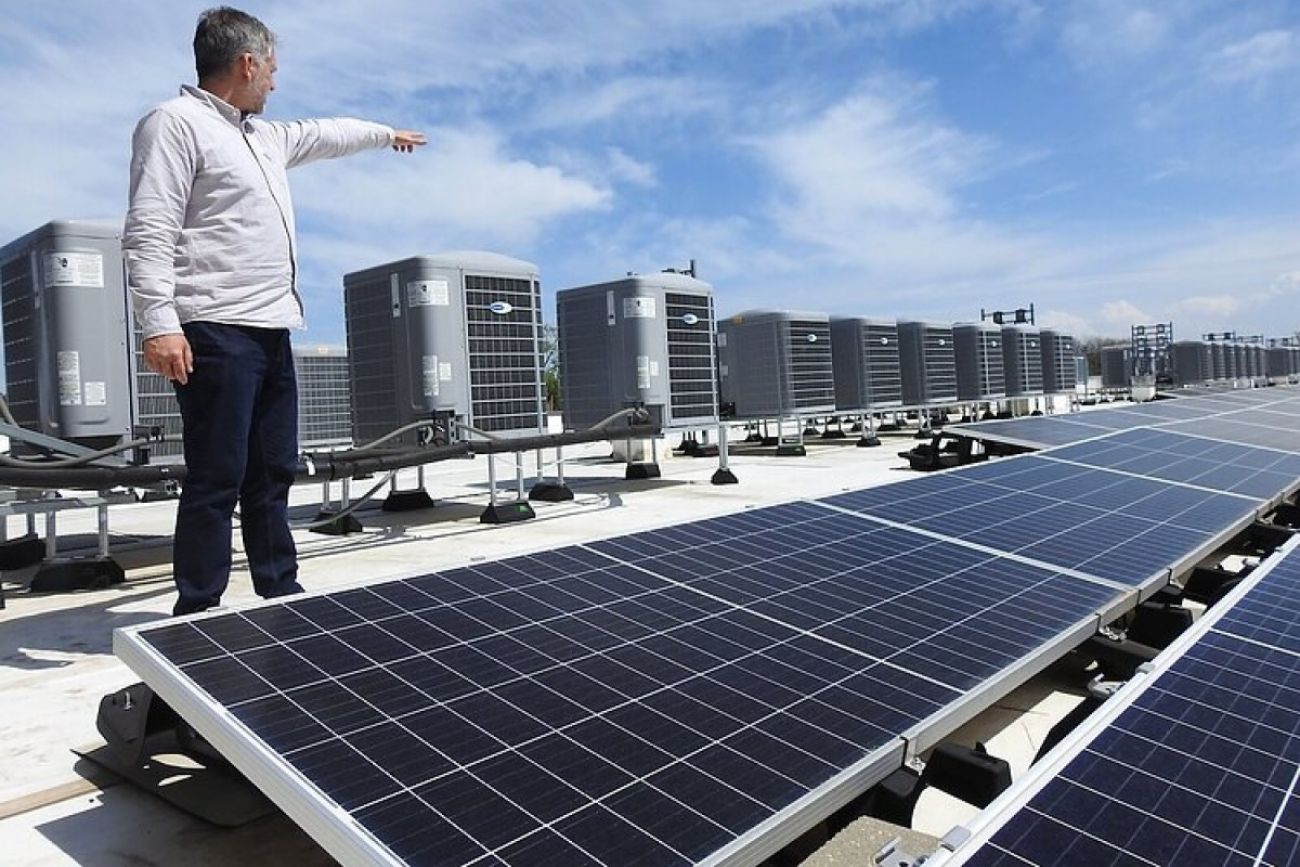 A man stands on a rooftop with solar panels in front of him and air conditioning units behind him and he points off into the distance