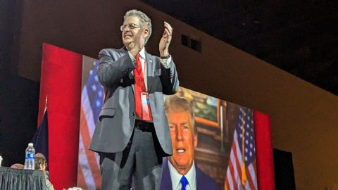 Matthew DePerno standing on a stage. A video of Donald Trump is behind him