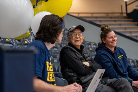 Sandy Vong sits in the bleachers, talking to two players.