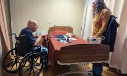 Howard Gregor in a wheelchair by a medical bed. A woman is standing on the other side of the bed.